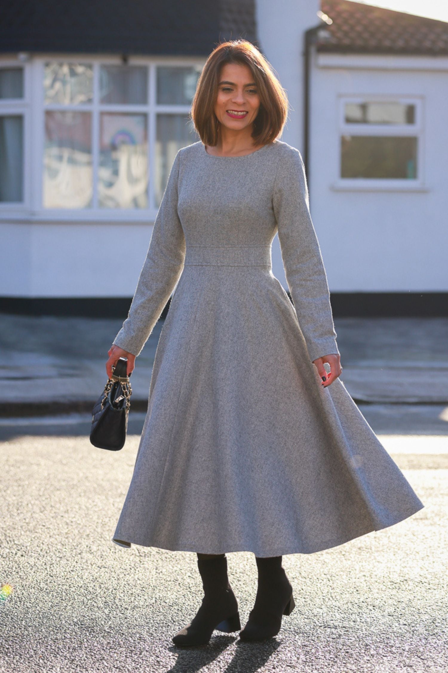 A woman standing on the sidewalk wearing a long sleeves wool gray dress with a round neckline, two side seam pockets, and black ankle boots.