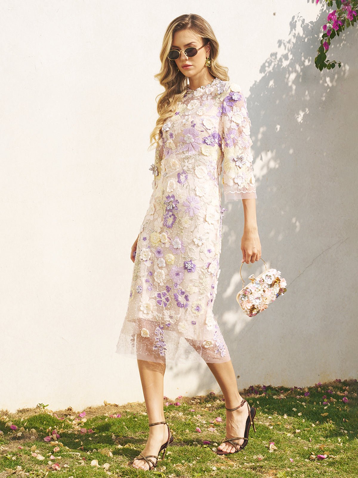 A woman in the Social Queen 3D Floral Embroidered Beaded Dress with a matching purse walks on grass beside a white wall in sunlight.
