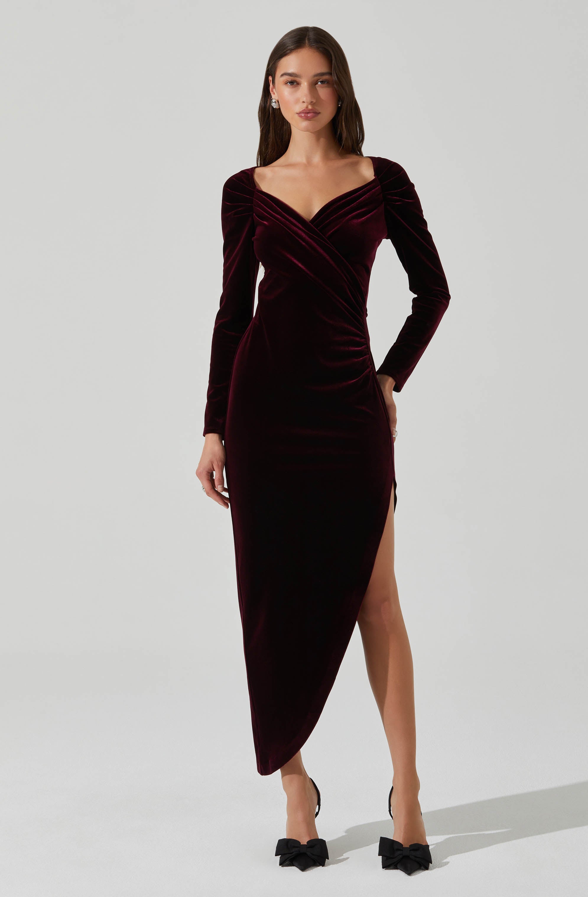 A woman is wearing a long, deep burgundy velvet dress with long sleeves and a fitted silhouette. The dress features a V-neckline, a draped, ruched bodice, and a high slit on one side, revealing her leg. She is paired with black high-heeled shoes adorned with large bows.