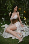Full body view of a silicone sex doll with black braided hair wearing a white and brown outfit sitting in a garden