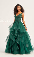 A woman stands against a plain background wearing the Ellie Wilde Tiered Long A-Line Prom Dress EW35119, featuring a sleeveless emerald green lace bodice with beaded details and a layered tulle skirt. "Ellie Wilde" appears on the left.