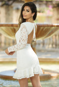 A woman in a white lace dress with long sleeves and a backless design stands gracefully, smiling over her shoulder. The background features a blurred fountain and greenery.