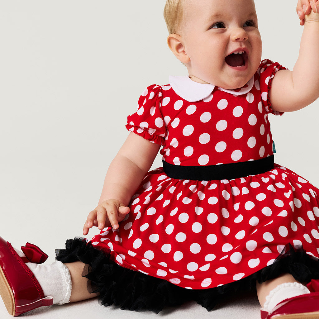 Baby wearing a red polka dot dress with a white collar and black belt on a light gray background