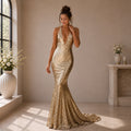 A woman stands in a sunlit interior wearing a floor-length gold sequin gown with a deep V neckline and a fitted mermaid silhouette with a slight train.